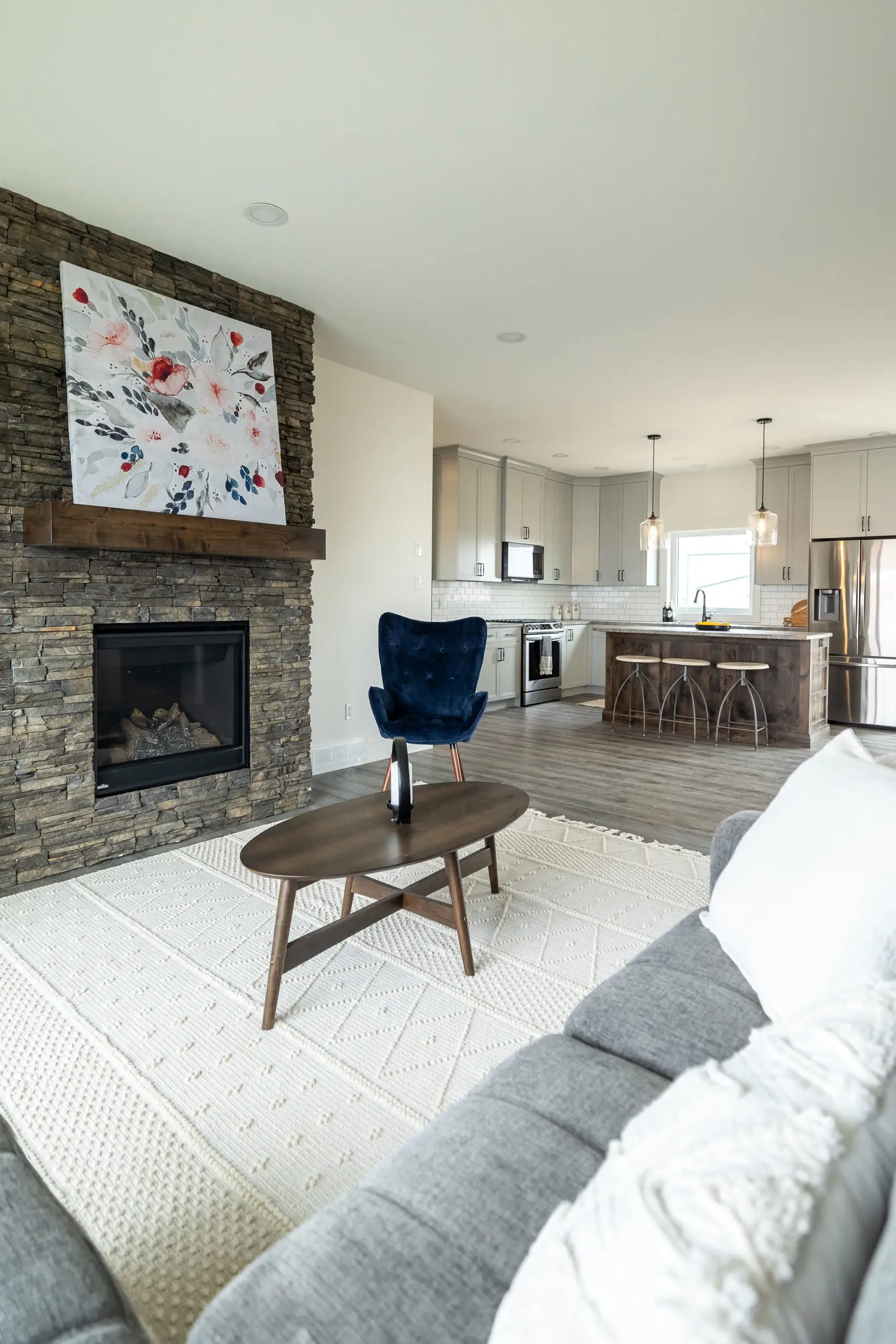 Modern living room with a stone fireplace and abstract art. Includes a gray couch, blue chair, kitchen in the background, and a cozy atmosphere.