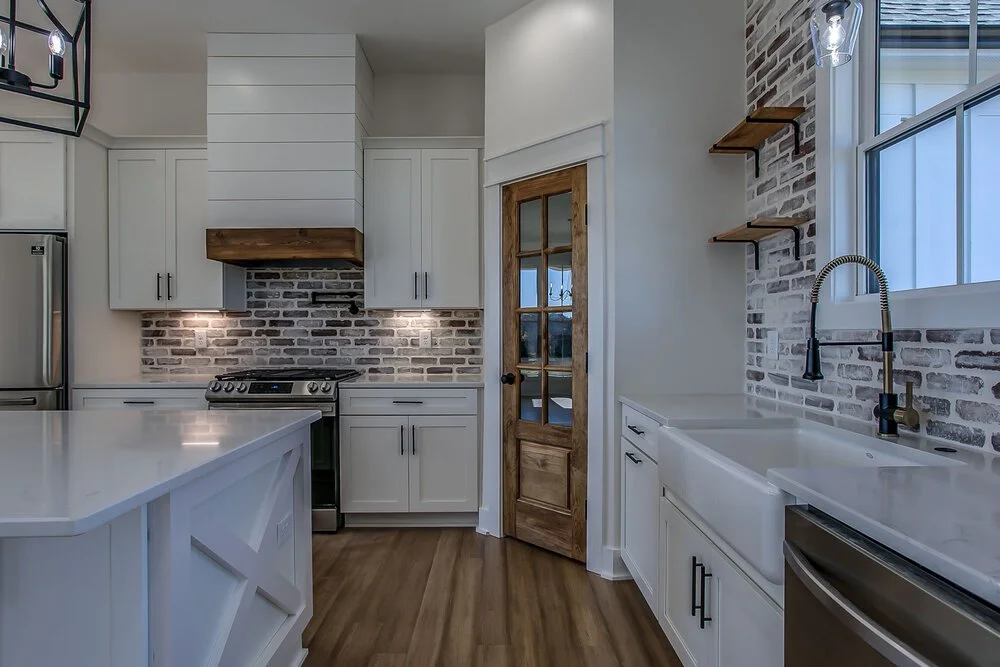A modern kitchen with white cabinets, brick backsplash, and wooden accents. It features a sleek stove, farmhouse sink, and a glass-door pantry.