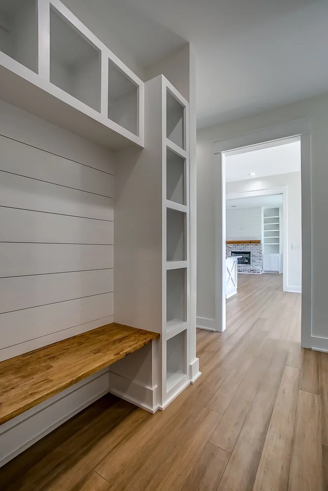 Bright hallway with wooden flooring, featuring built-in white shelves and a wooden bench. The area leads to a cozy room with a fireplace, exuding warmth.