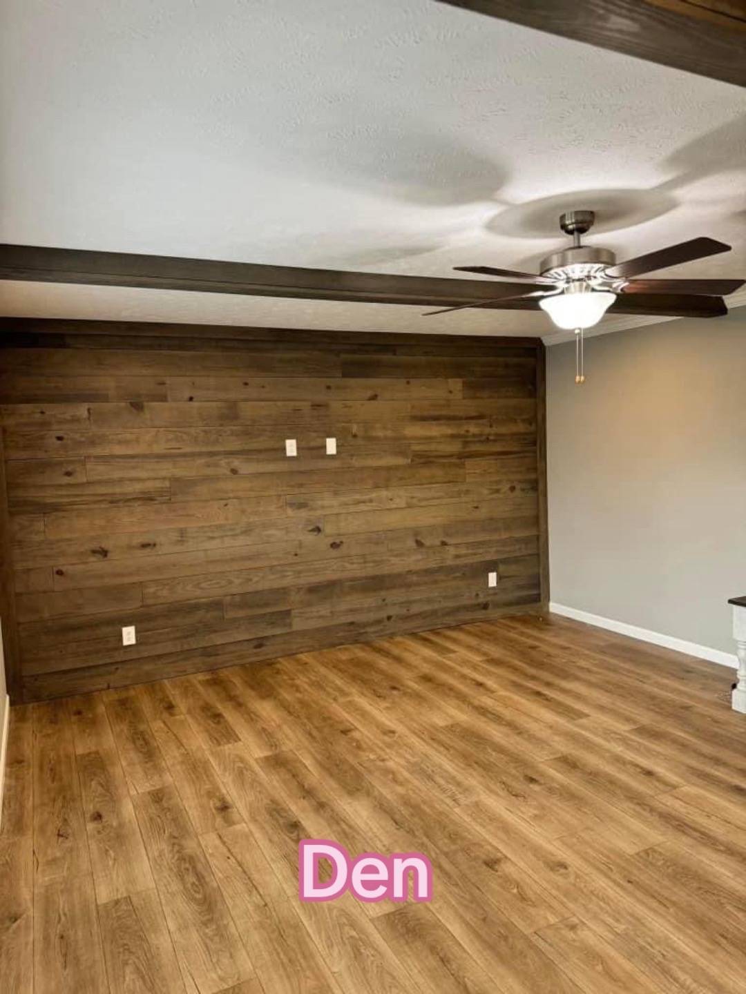 A room labeled "Den" with wood panel flooring and walls. A ceiling fan with lights hangs from the white ceiling. The atmosphere is warm and inviting.
