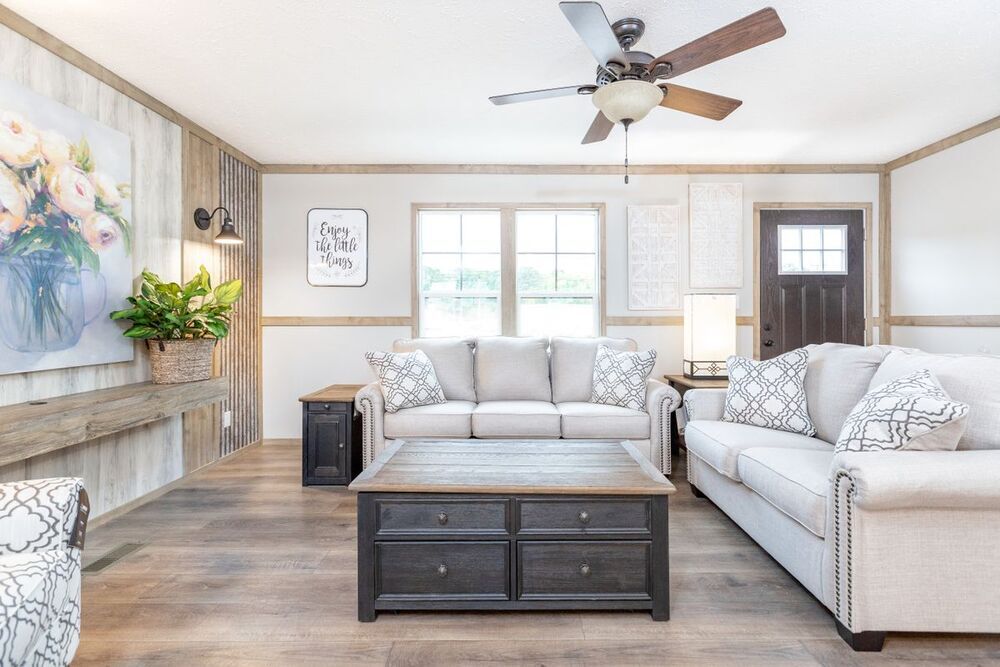 Charming living room with light gray sofas, patterned cushions, dark wood coffee table, ceiling fan, and floral wall art. Bright, cozy, and inviting atmosphere.