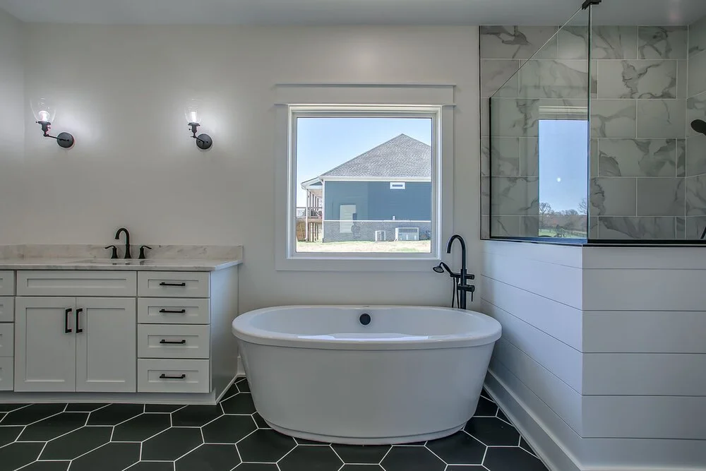 Modern bathroom with oval bathtub, black hexagonal tiles, and white vanity with black fixtures. Large window and glass-enclosed shower on the right.