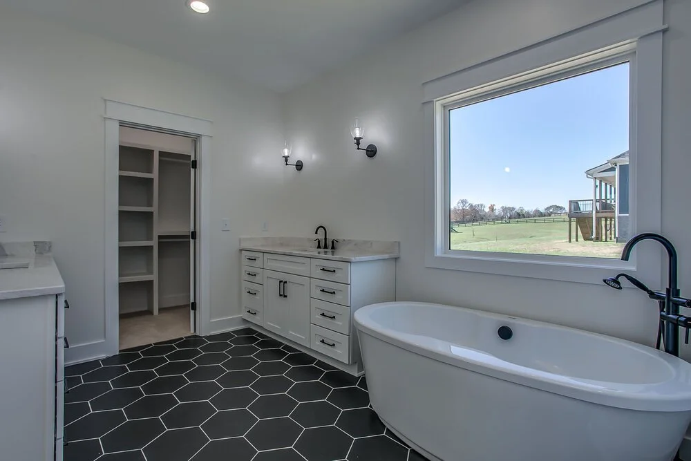 Modern bathroom with a freestanding tub, marble vanity, and black hexagonal floor tiles. Large window overlooks a sunny landscape, creating a serene atmosphere.