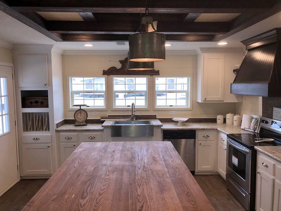 Rustic kitchen with a large wooden island, farmhouse sink, and dark beamed ceiling. White cabinets, metal pendant light, and a vintage scale enhance charm.