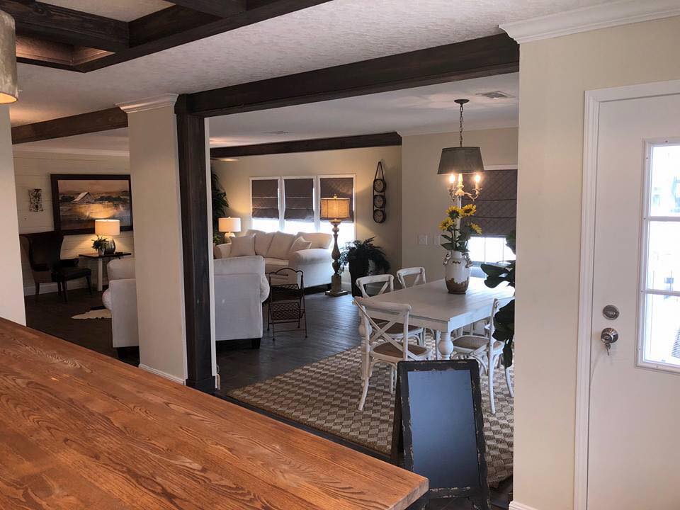 Open-concept living area with a cozy white sofa, wooden dining table, dark wooden beams, and warm lighting. The decor is modern and inviting.