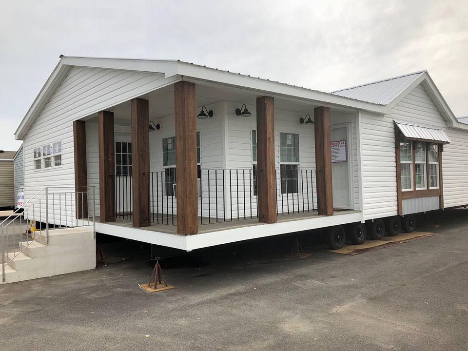 White mobile home with a gabled roof and wooden porch posts on wheels. It features a small porch with railings and several windows, set on a paved area.