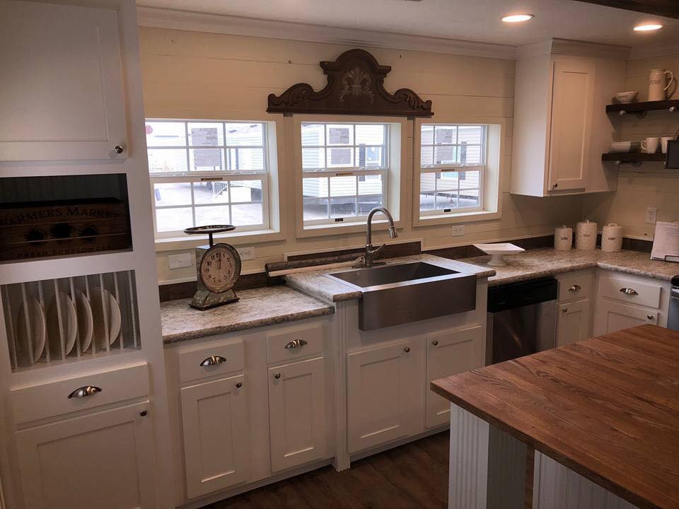 Rustic kitchen with white cabinets, farmhouse sink, and wooden countertops. Vintage scale on counter. Large windows create a cozy, warm ambiance.