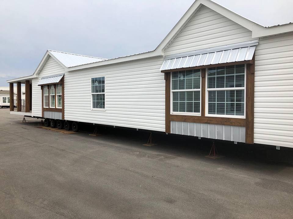 A white manufactured home with brown trim and large windows sits on blocks in an outdoor lot. The setting is overcast, conveying a neutral tone.