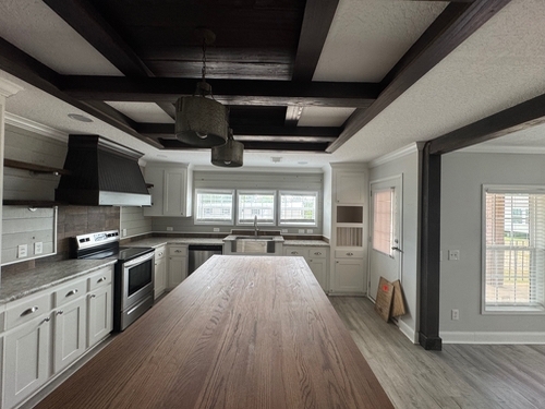 Spacious kitchen with modern farmhouse style; features a large wooden island, white cabinets, stainless steel appliances, and exposed beams. Natural light floods in through windows, creating a cozy ambiance.