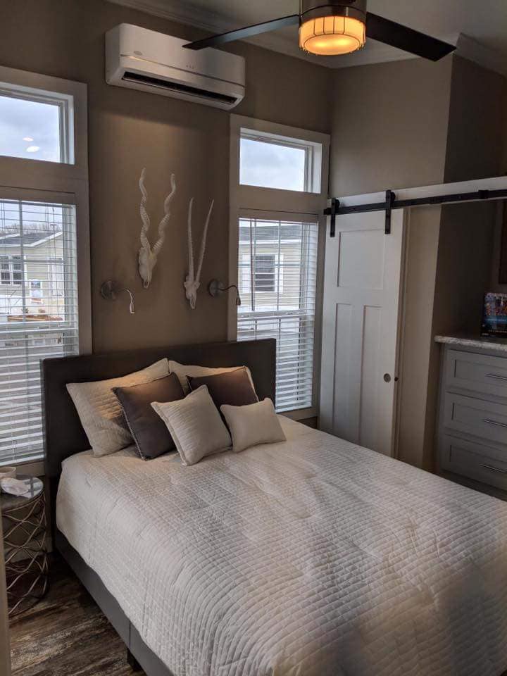 A cozy bedroom with a neatly made bed, neutral-toned pillows, white quilt, and decorative wall hangings. Soft lighting and clean lines create a calming atmosphere.