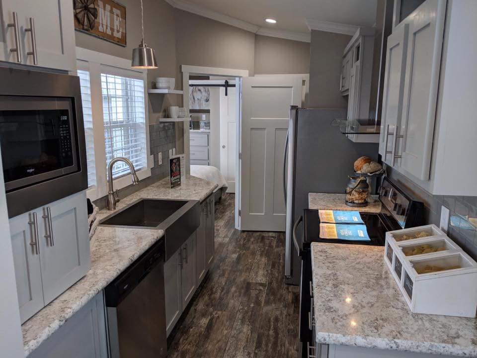 Sleek modern kitchen with gray cabinets, granite countertops, stainless steel appliances, and dark wood flooring. Sunlight through large windows adds warmth.