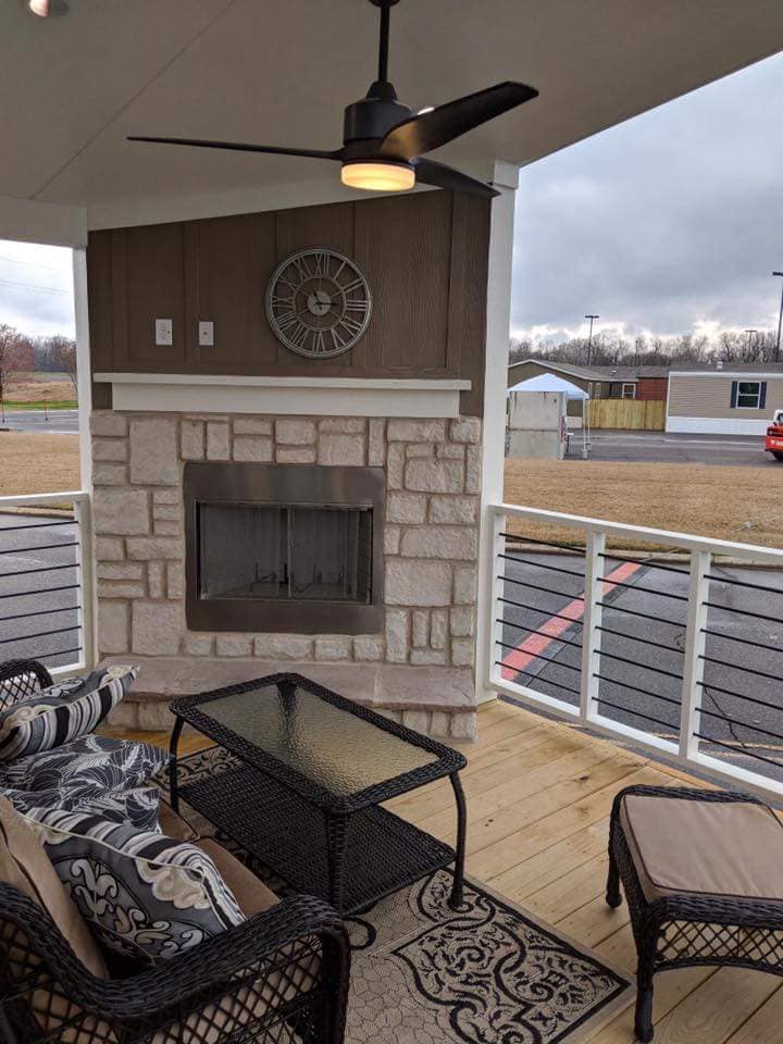 Cozy porch with stone fireplace, wicker furniture, patterned cushions, and glass table. Ceiling fan and wall clock above. Overcast sky outside.