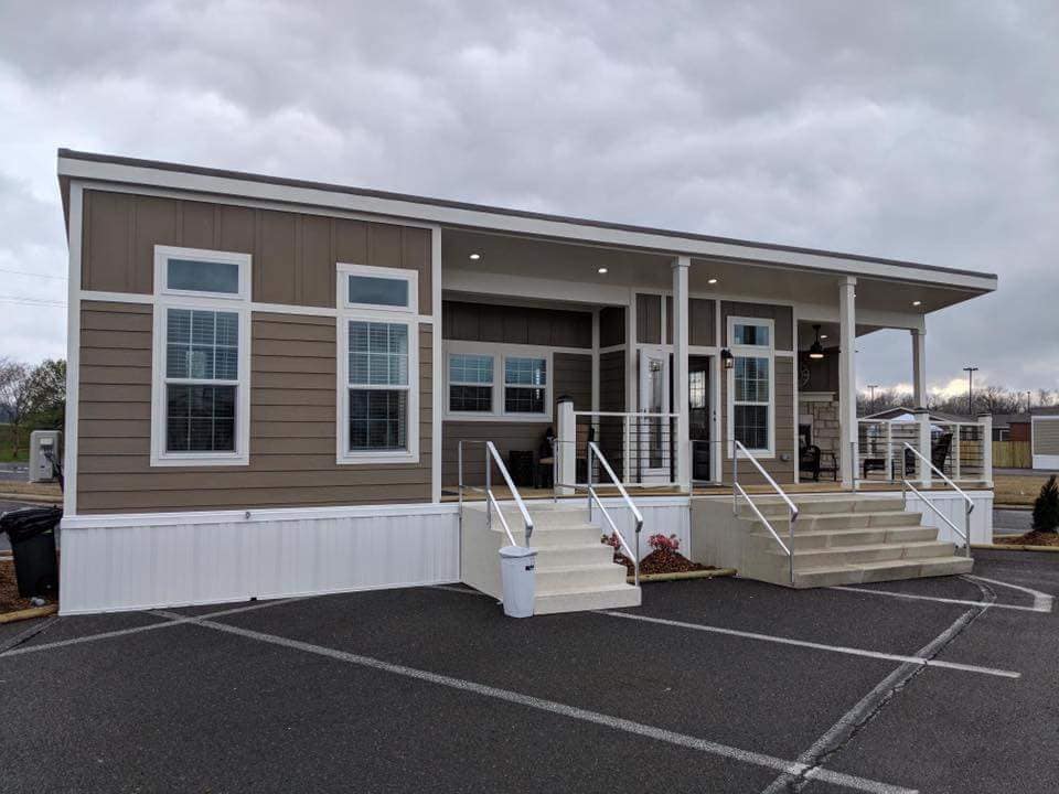 A modern, tan modular home with large windows and white trim. It features two sets of stairs with railings and is set against a cloudy sky.