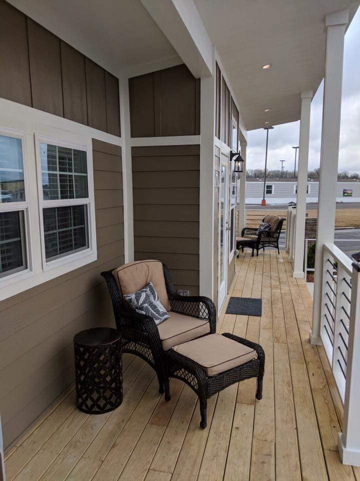 Covered porch with wooden floorboards, featuring a wicker chair with beige cushions and a matching ottoman. Another chair is visible further down. Cozy and inviting atmosphere.