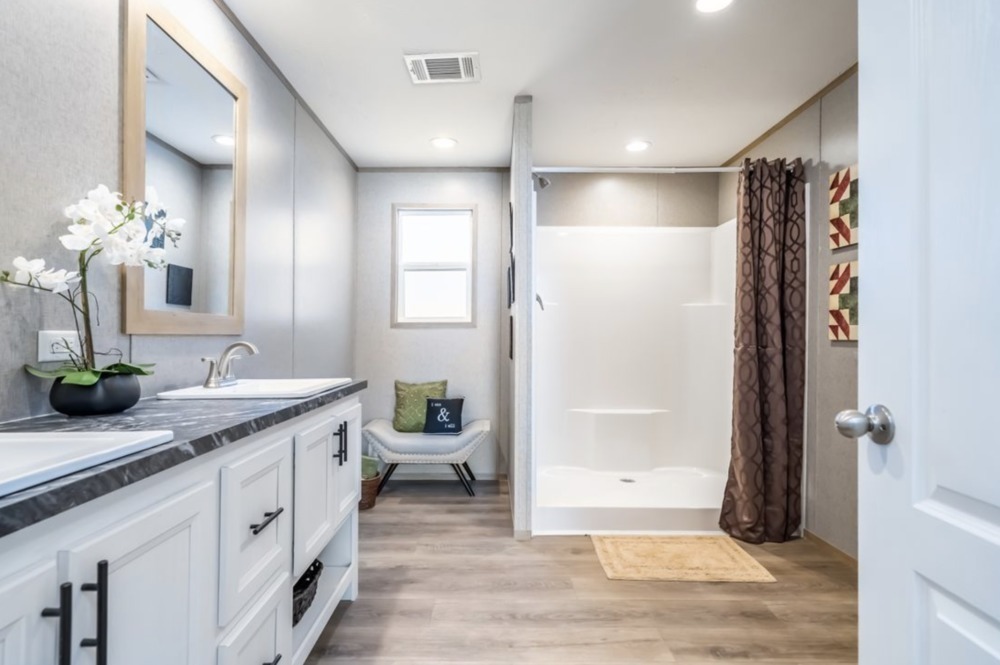 Modern bathroom with white cabinets, dual sinks, and orchid decor. A shower stall with a brown curtain is on the right, and a chair with cushions is near the window. Warm, inviting ambiance.