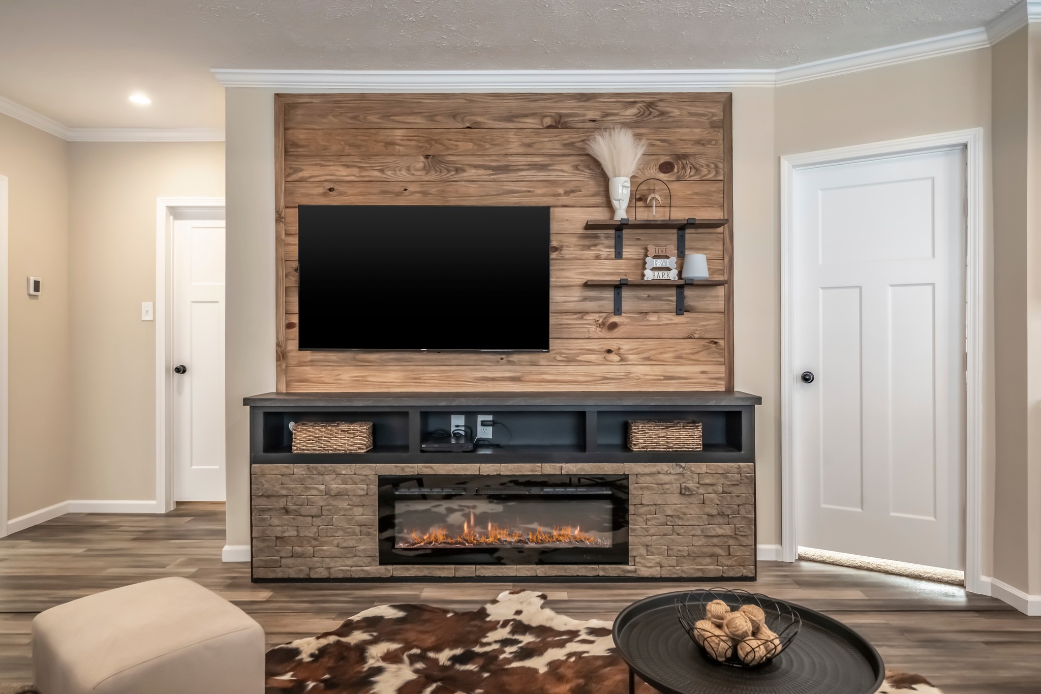 Cozy living room with a wood accent wall featuring a mounted TV and shelves. Below is a stone-faced electric fireplace. A round table with decor sits on a cowhide rug.