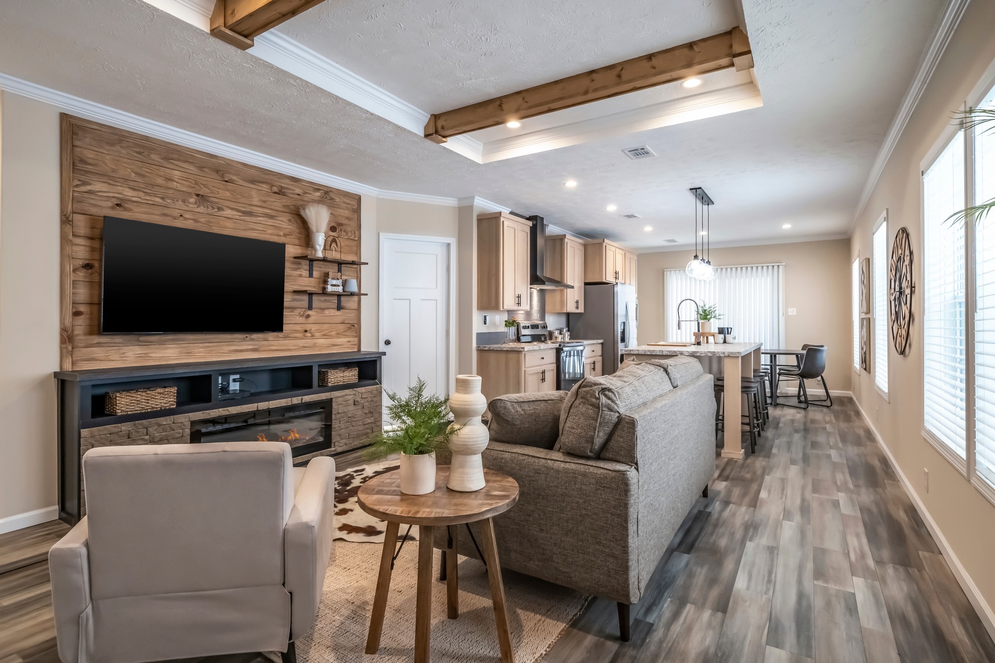 Modern open-concept living room and kitchen with wood accents. Features cozy seating, flat-screen TV, and dining area with hanging lights. Bright and inviting atmosphere.