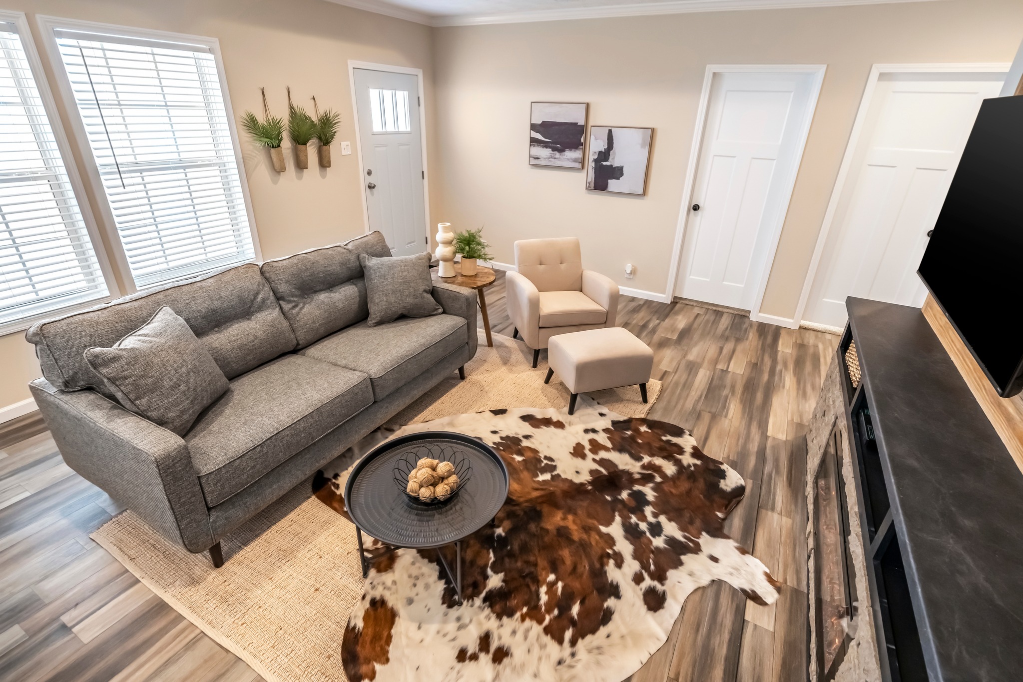 Stylish living room with gray sofa, beige armchair, and cowhide rug on wooden floor. Walls are cream-colored with modern art and potted plants. Warm, cozy atmosphere.