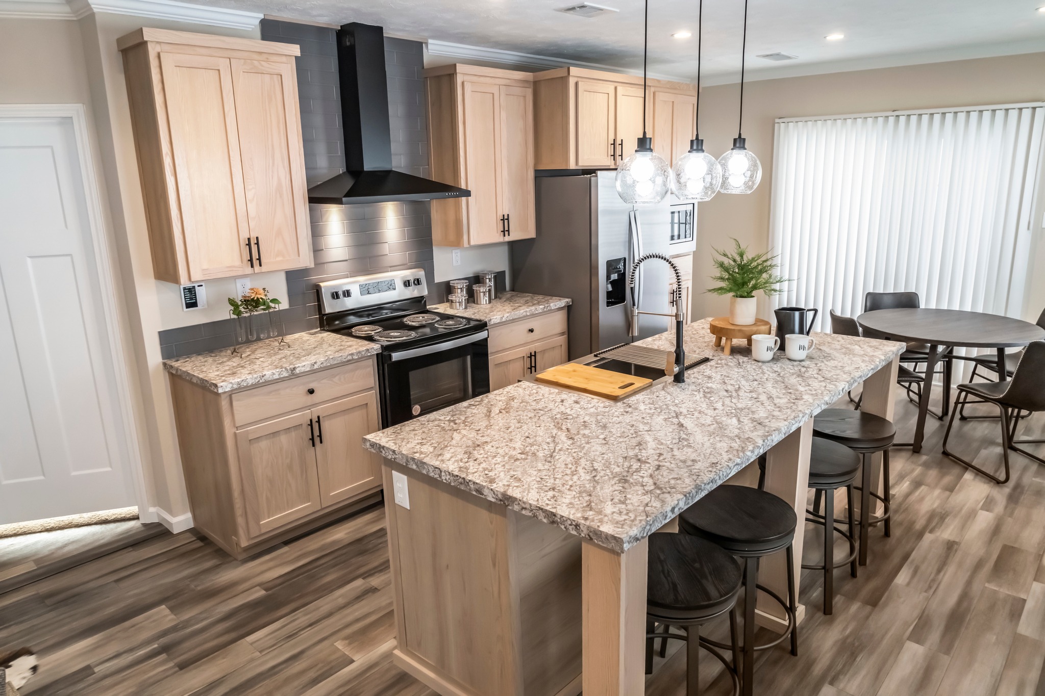 Modern kitchen with light wood cabinets, granite countertops, and a central island featuring bar stools and hanging lights. Cozy dining area in the background.