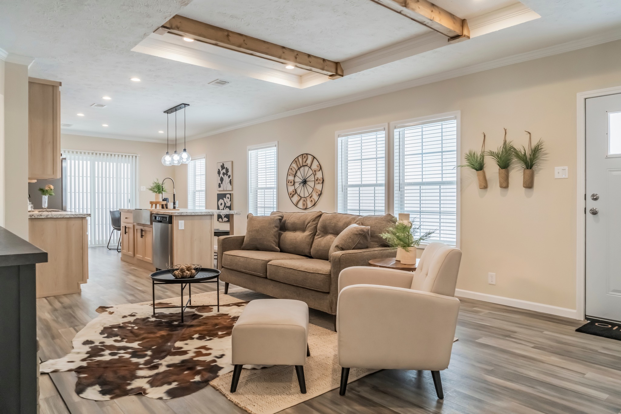 Modern living room with open kitchen. Features a beige sofa, cowhide rug, and large wall clock. Airy feel with light streaming through windows.