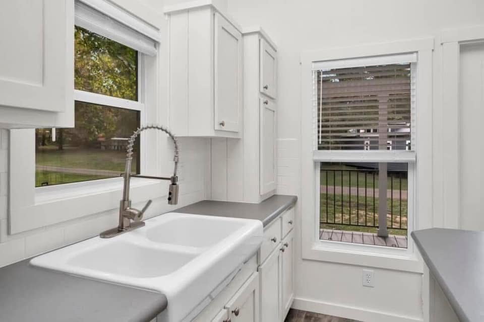 Bright kitchen with white cabinets, a farmhouse sink, and stainless steel faucet. Large window shows a green outdoor view, creating a fresh ambiance.