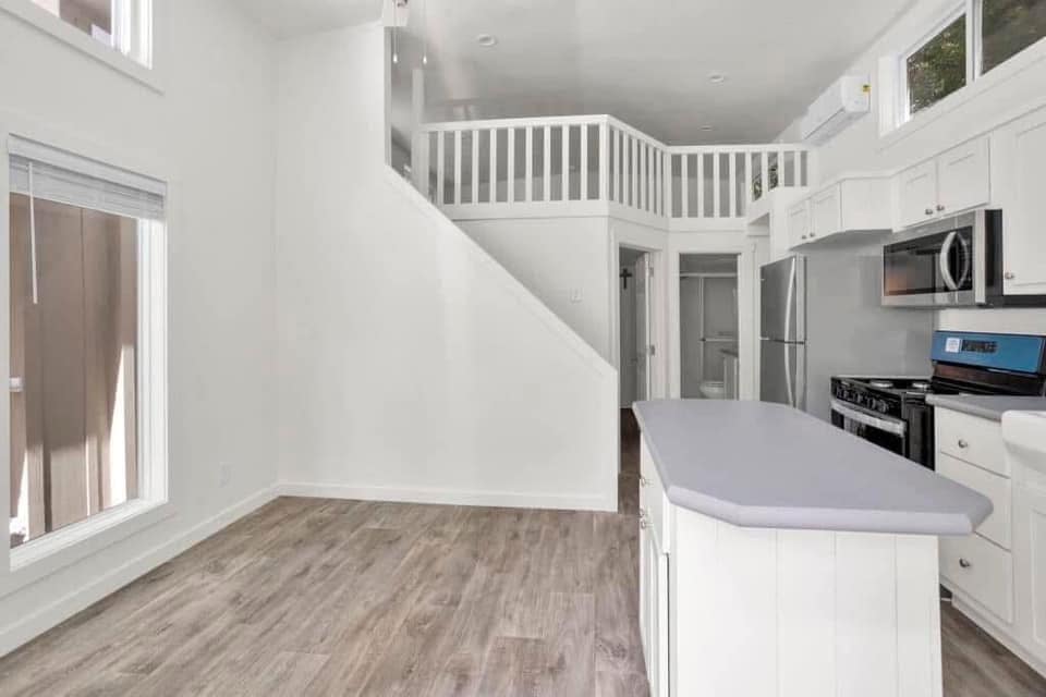 Bright, modern kitchen with white cabinetry, stainless steel appliances, and light wood flooring. Loft area with railing above staircase adds depth.