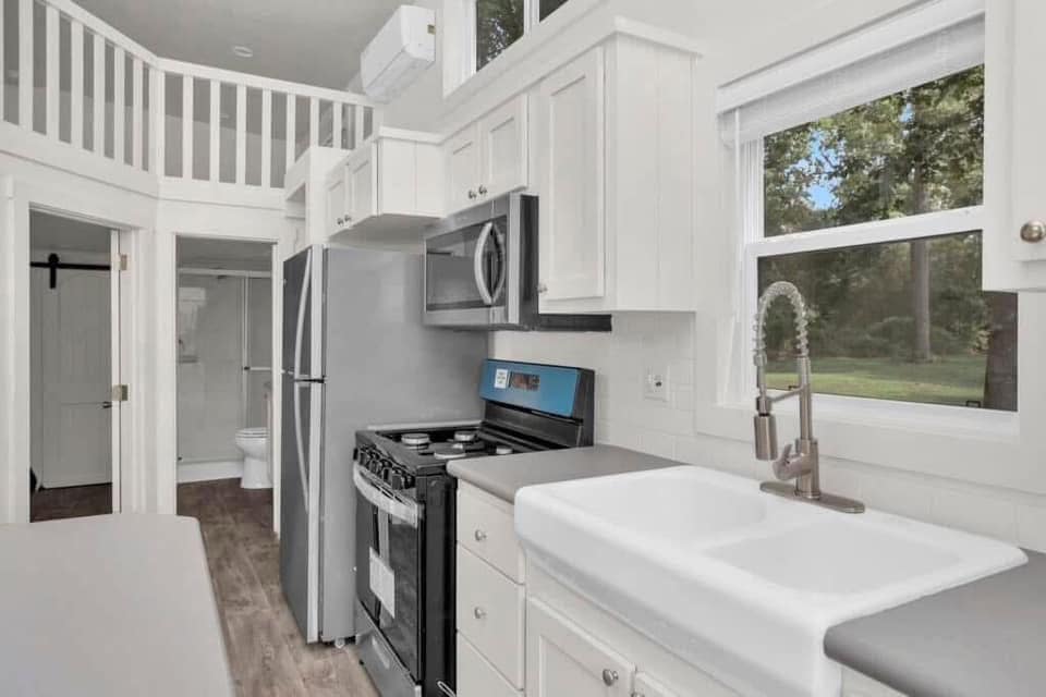 Bright modern tiny house kitchen featuring white cabinets, stainless steel appliances, and a large farmhouse sink. Sunlit window with garden view.