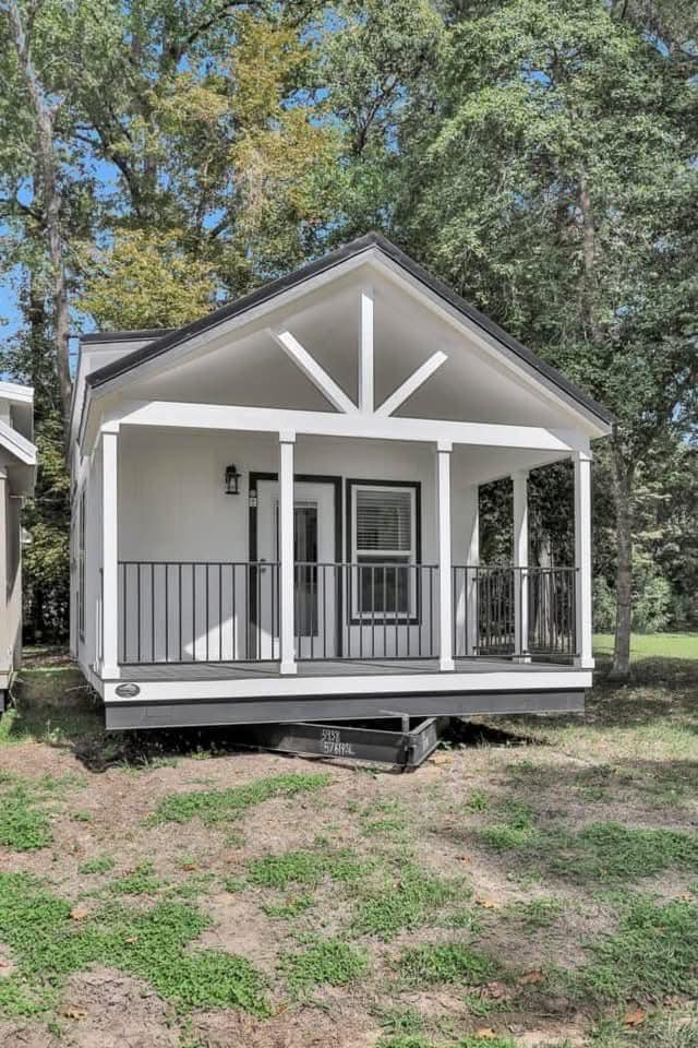 Small white house with a black railing front porch, set in a grassy area against a backdrop of tall trees. The scene conveys a peaceful, cozy atmosphere.