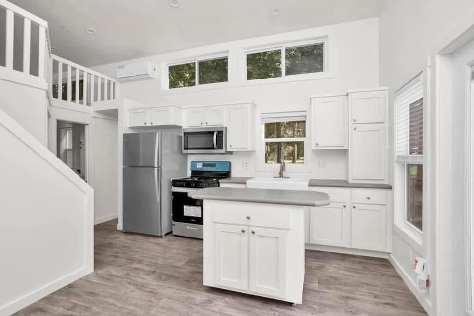 Bright, modern kitchen with white cabinets and stainless steel appliances. A compact gray island sits centrally under high windows, exuding a clean, airy feel.