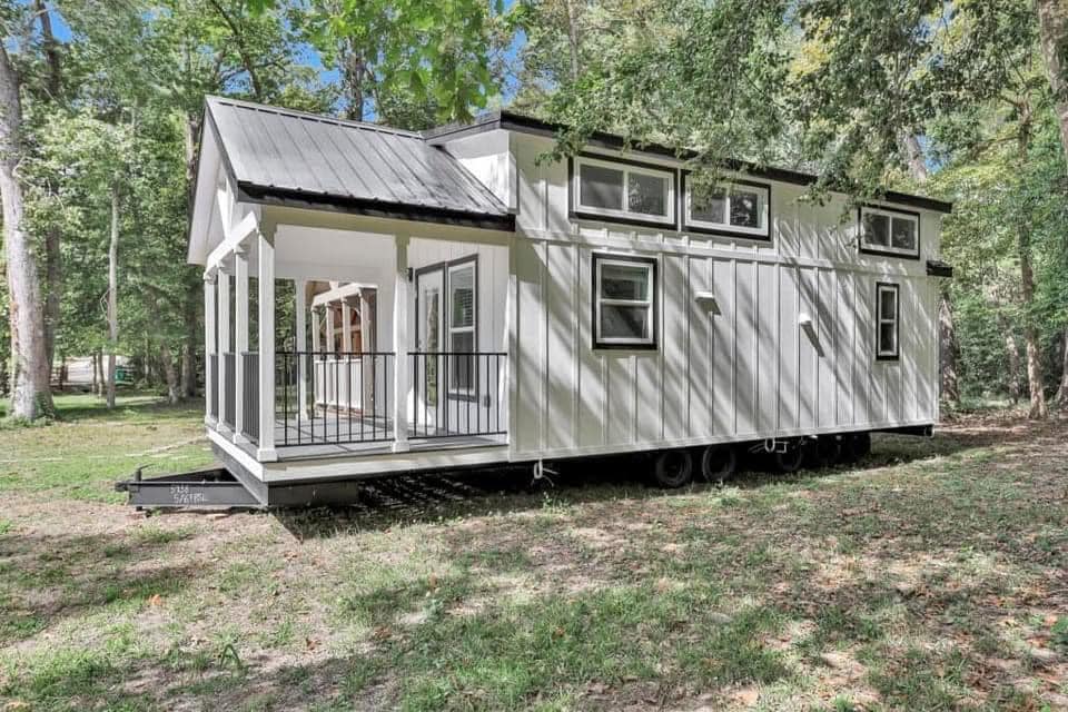 A white tiny house with a black metal roof sits on wheels in a wooded area. It features multiple windows and a small porch, conveying a cozy, modern vibe.