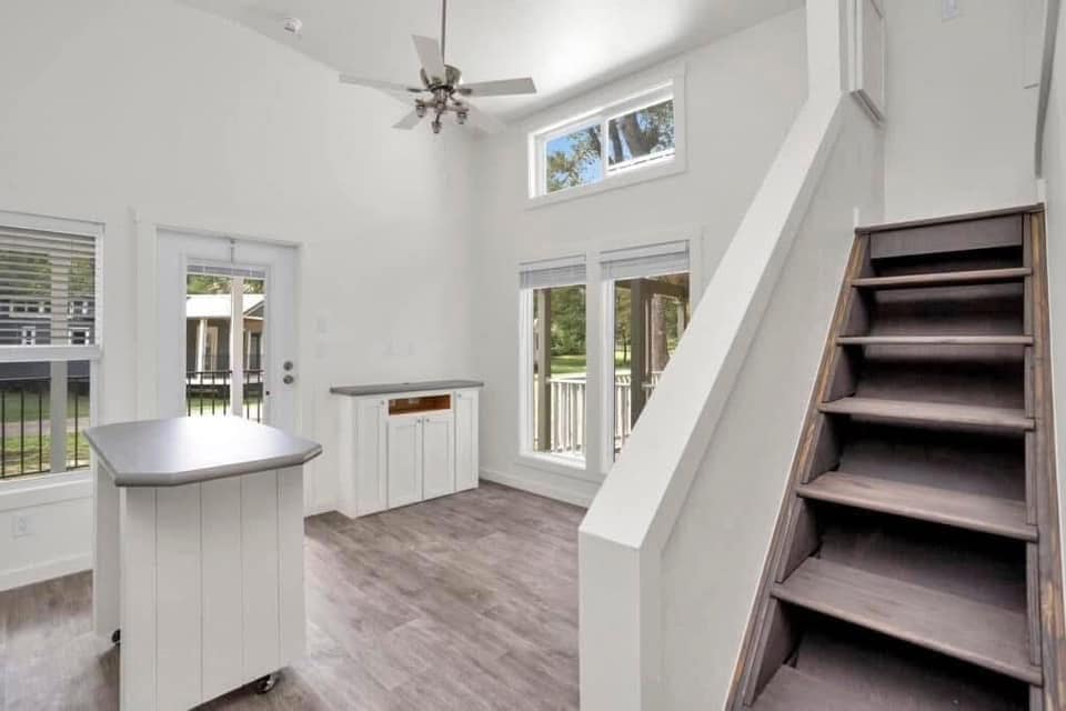 Modern, bright room with white walls, high ceiling, and large windows. Features dark wood floors, a ceiling fan, staircase, and a kitchen island.