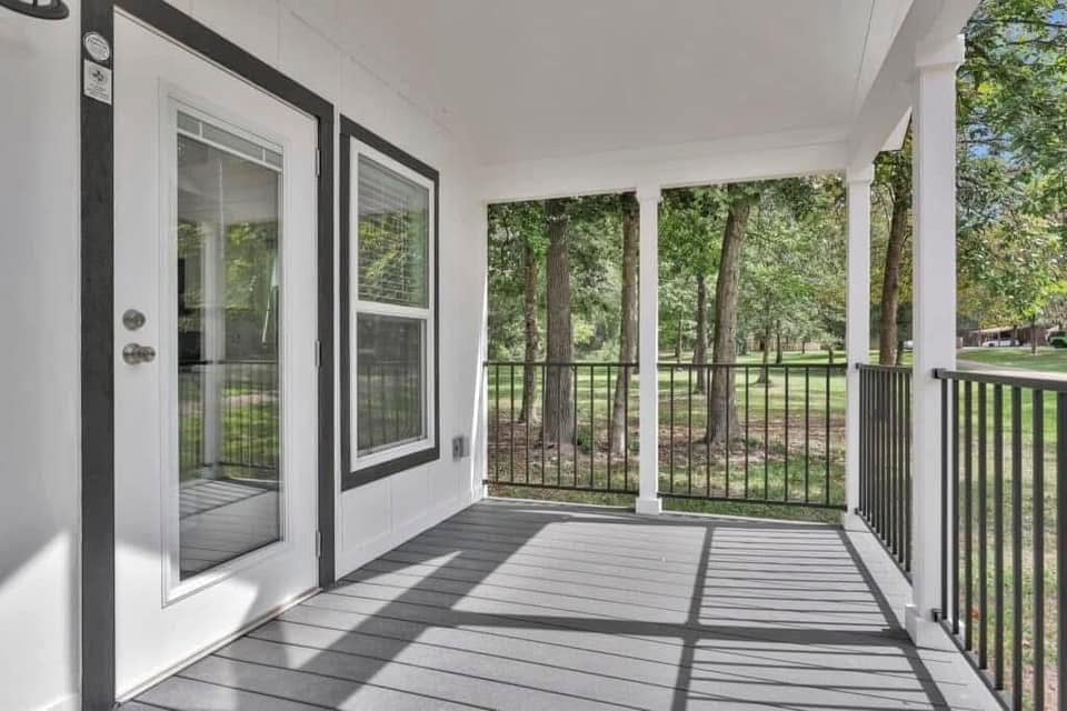 Covered porch with wooden flooring, white trim, and a glass door. Black railing overlooks a grassy area with trees, creating a serene, inviting atmosphere.