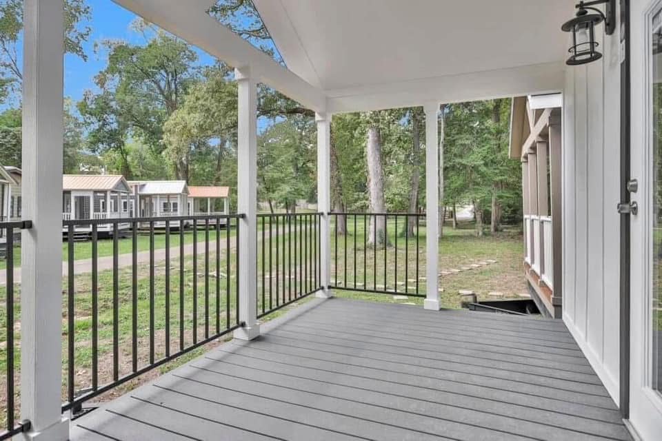 Covered porch with black railing overlooking a lush green backyard and distant cottages on a sunny day, conveying a peaceful and inviting atmosphere.