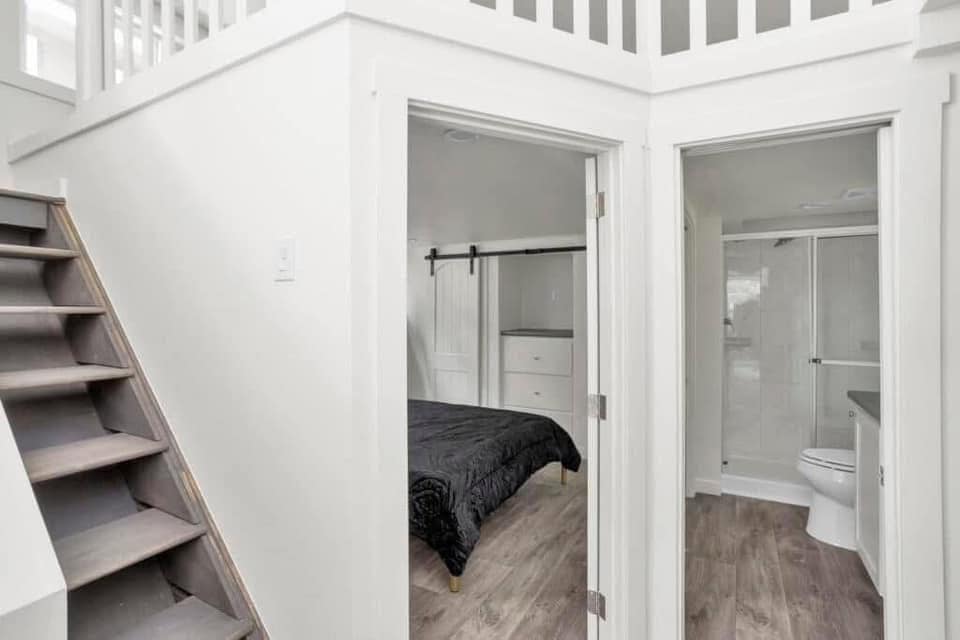 Cozy tiny house interior with a wooden staircase leading up. Below, a bedroom with a black quilt and a small bathroom featuring a glass shower door.