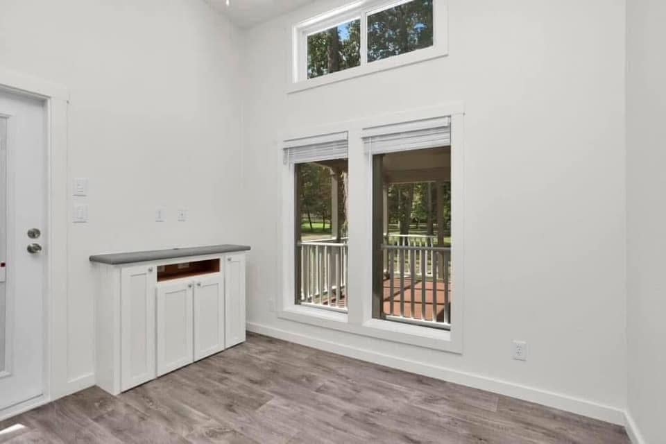 Bright room with white walls, wood flooring, and large windows revealing a green outdoor view. A small white cabinet sits against the left wall.