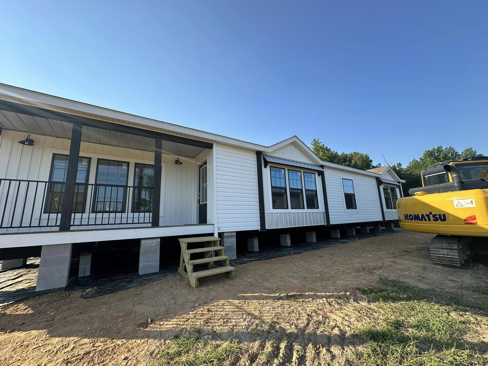 Single-story white manufactured home with black trim, elevated on stilts. A yellow excavator is nearby, under a clear blue sky.