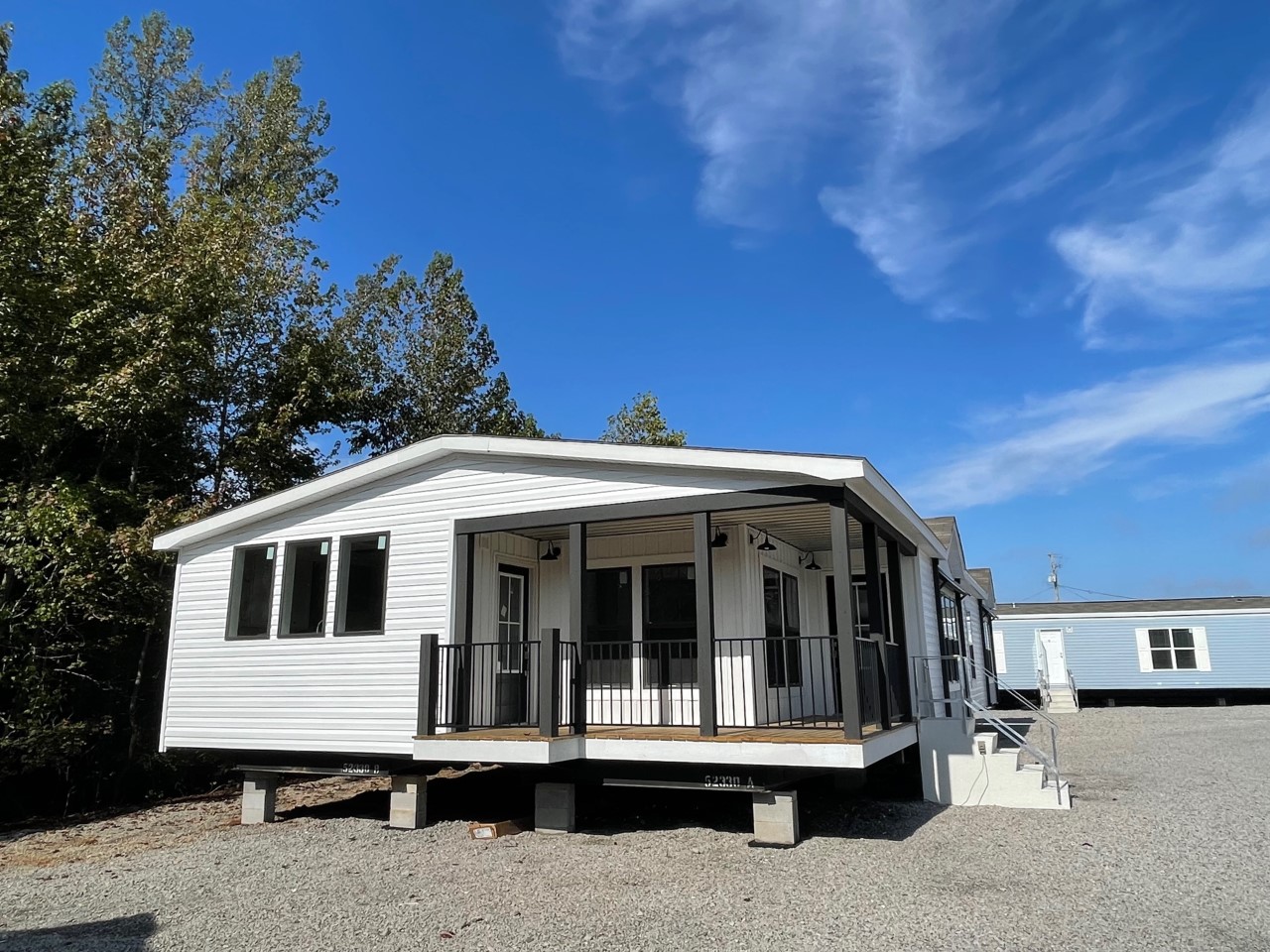 A modern white modular home with a covered porch and black railings sits on a gravel lot under a clear blue sky, surrounded by trees.