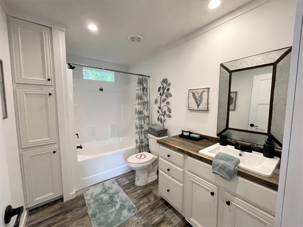 A modern bathroom with white cabinets, a wooden countertop, and a sink beneath a framed mirror. The shower has a floral curtain, and a green rug lies on the wooden floor. A wall decor and towels add a cozy touch.