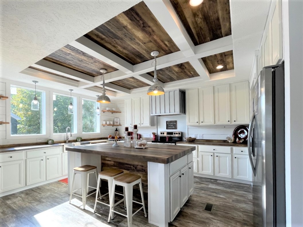 Spacious kitchen with rustic wooden ceiling panels, white cabinets, and an island with stools. Large windows and industrial pendant lights add brightness.