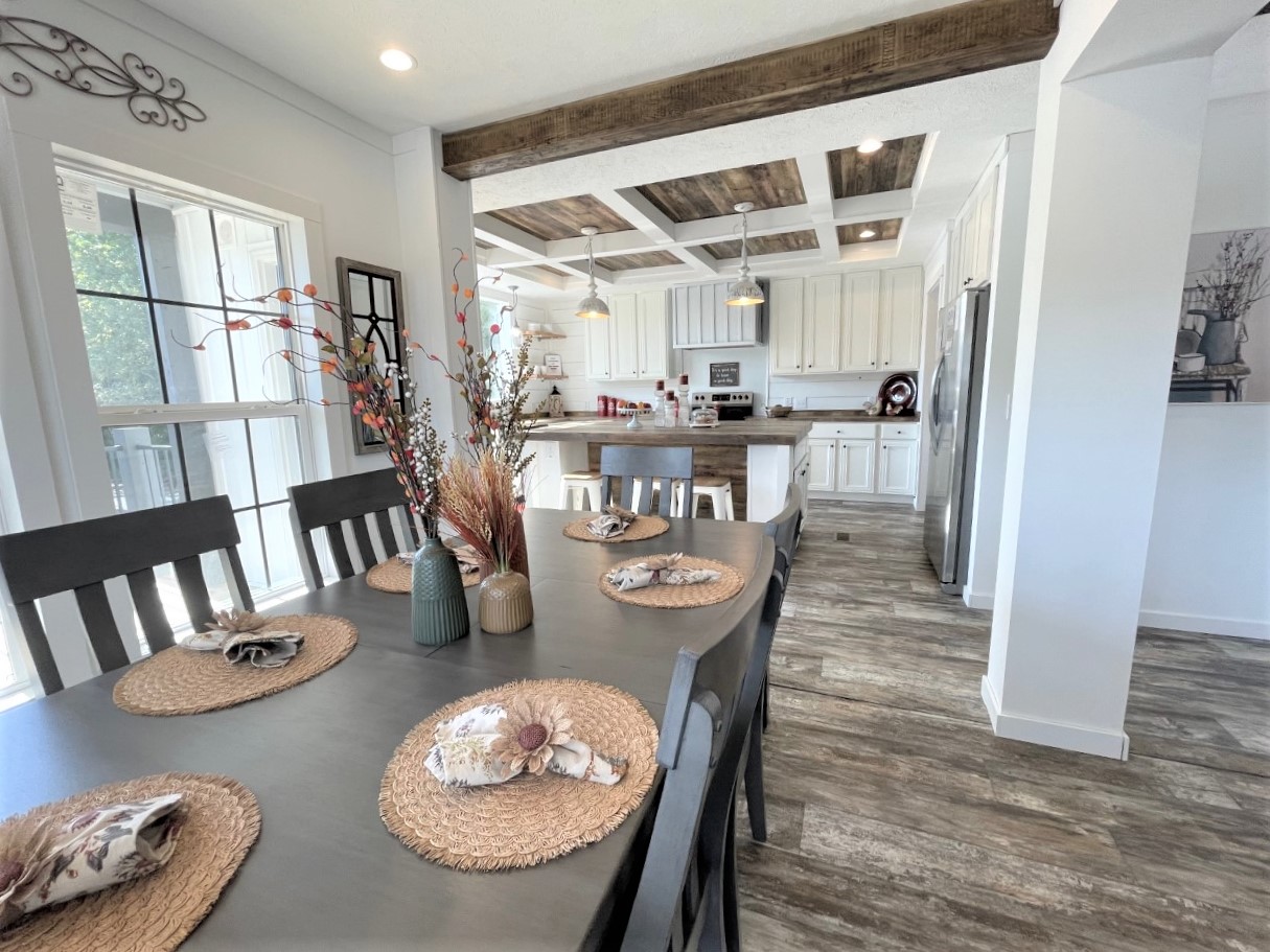Cozy dining area with a dark wooden table, fall-themed centerpieces, and woven placemats. Bright kitchen in the background with rustic decor.