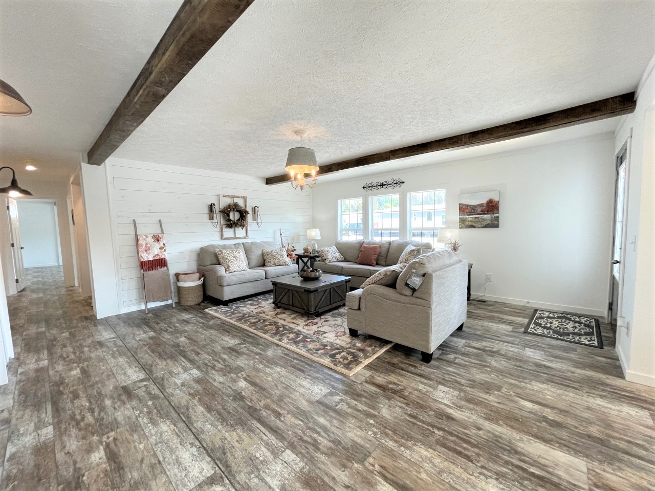 Spacious living room with rustic beams, a white shiplap wall, and a cozy gray sectional sofa. Features decorative cushions, a central dark wood coffee table, and a warm, inviting ambiance.