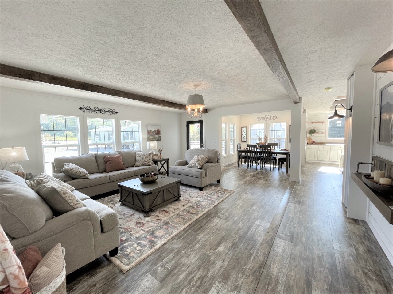 Spacious living room with gray sofas, wooden floors, and ceiling beams. Bright dining area and kitchen in background, creating an inviting, cozy atmosphere.