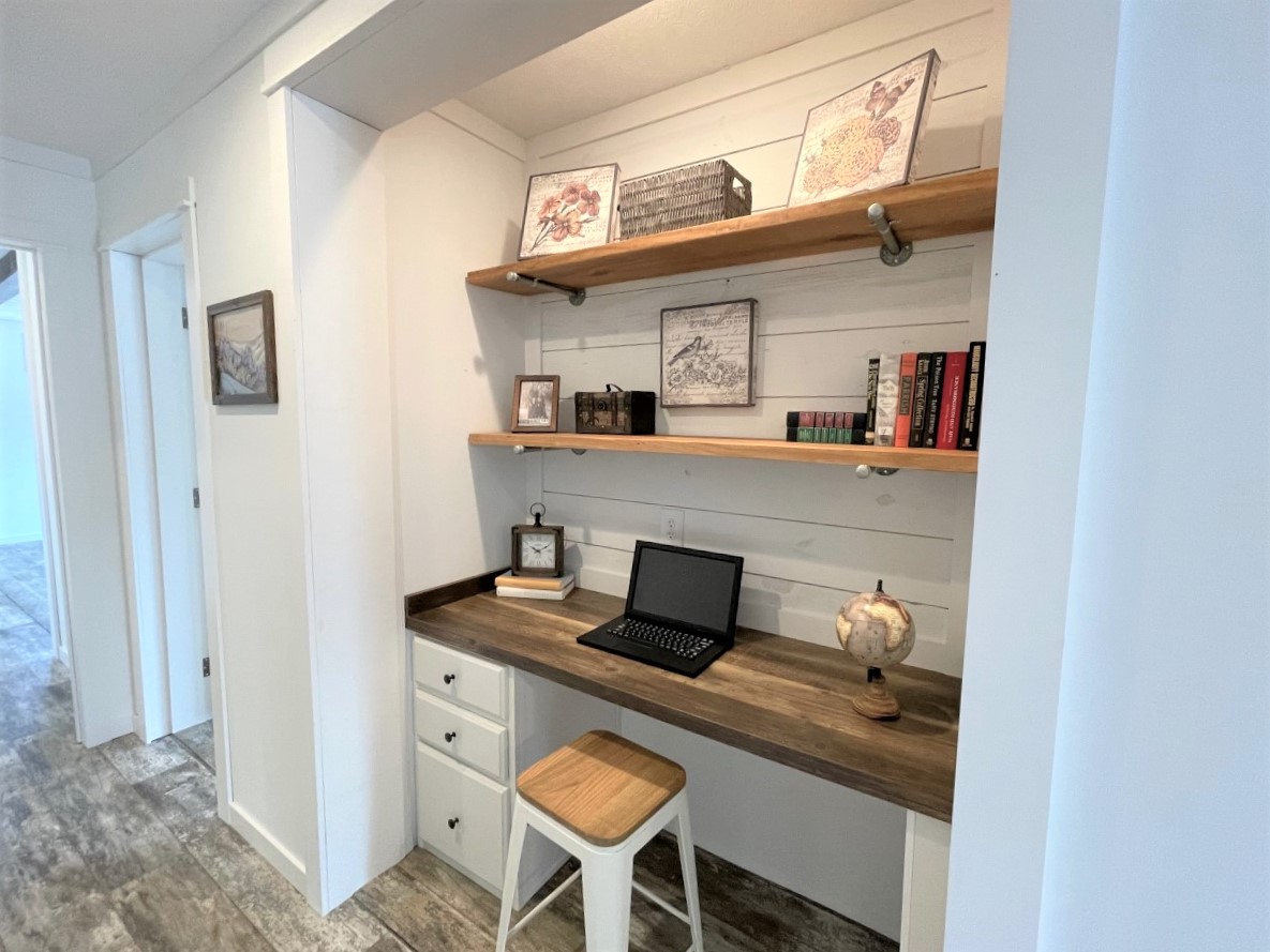 Cozy home office nook with a wooden desk, laptop, and small globe. Two wall shelves above hold books and decorative items, creating a warm, inviting workspace.