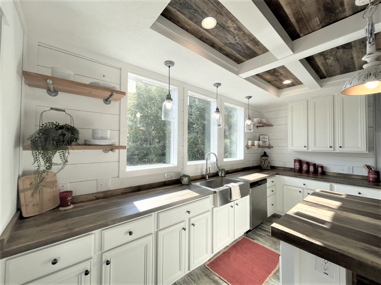 Bright farmhouse kitchen with white cabinets, rustic wood countertops, and pendant lights. Large windows illuminate open shelves with plants, dishes, and decor. Cozy and inviting atmosphere.