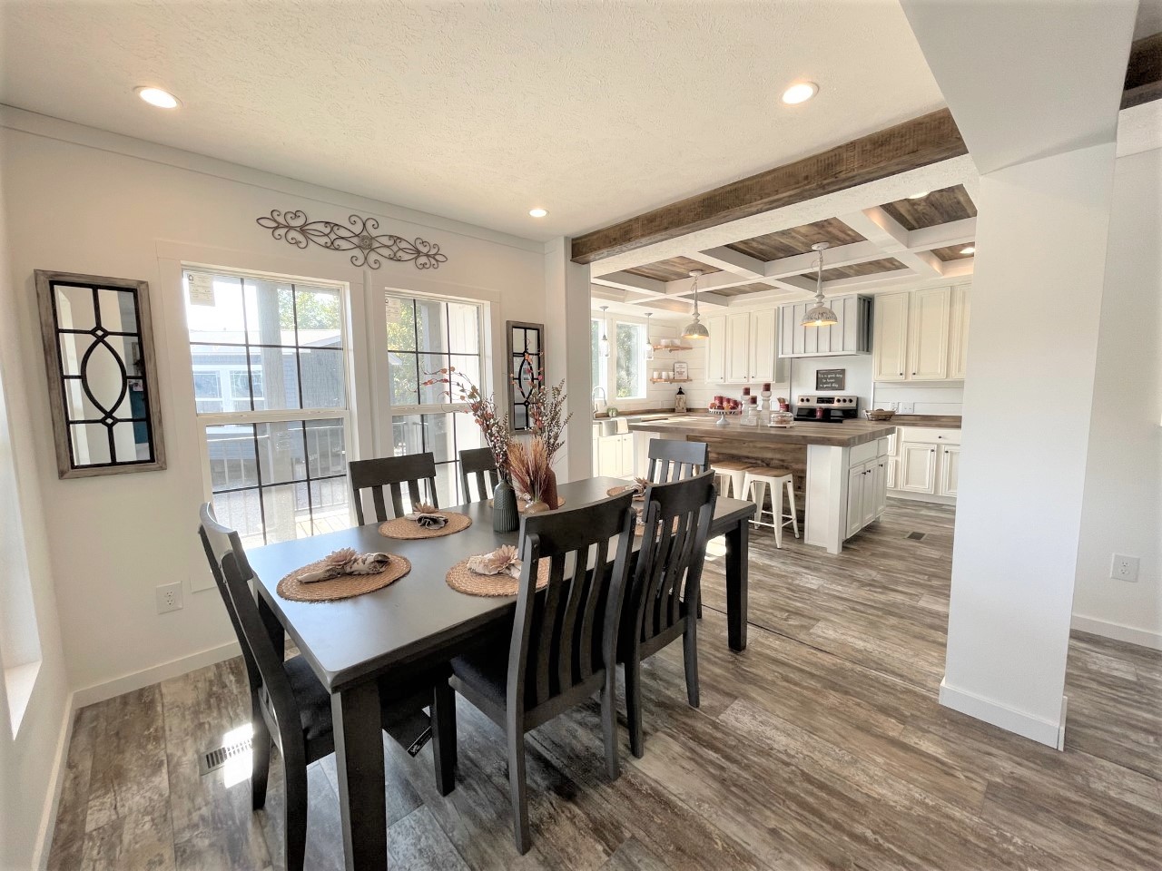Bright dining area with a wooden table for six, rustic decor, chandelier, and large windows. Open kitchen with island and modern appliances.