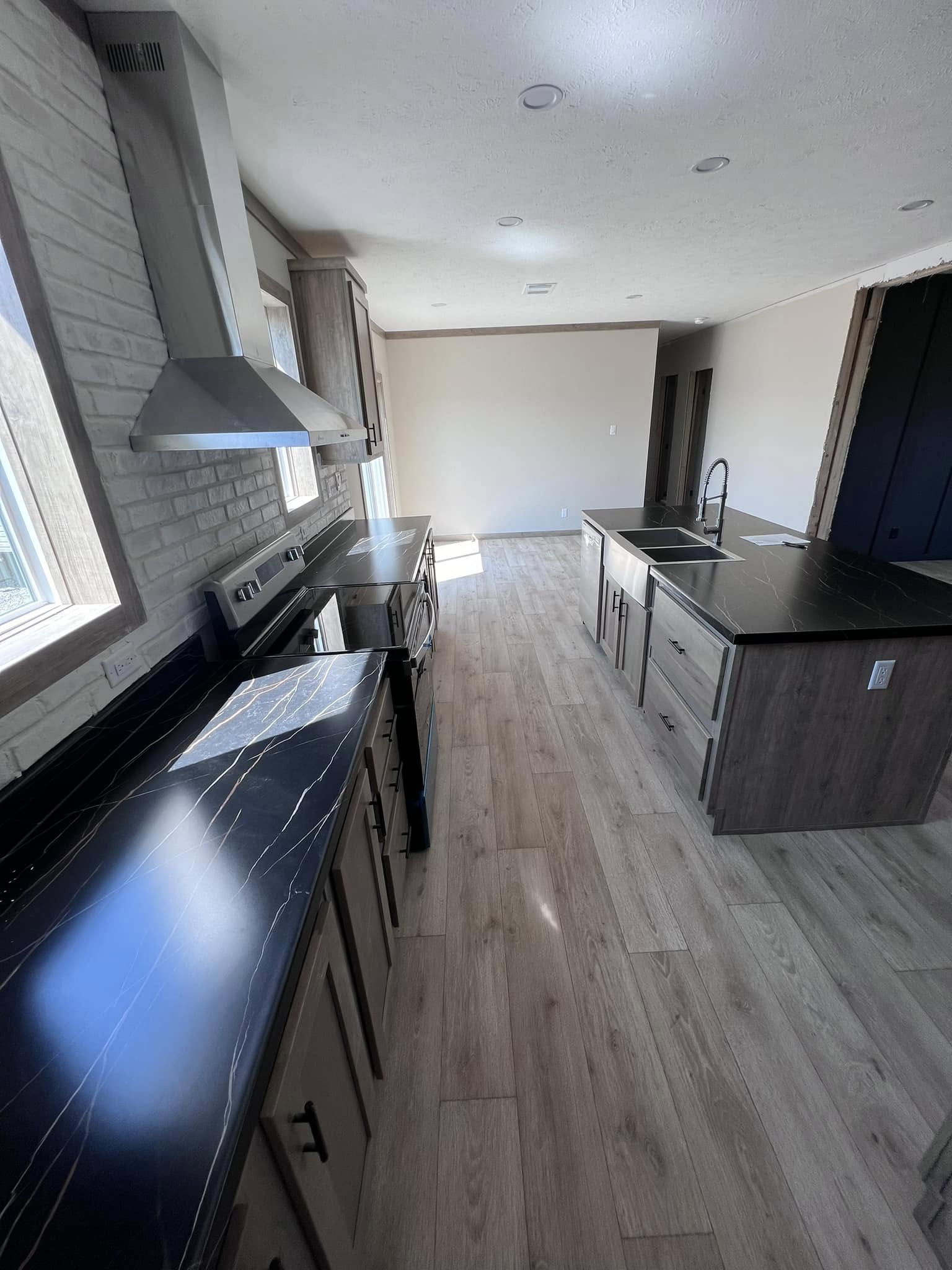 Modern kitchen with gray wood cabinets, black marble countertops, and a stainless steel range hood. Sunlight brightens the light wood floor.