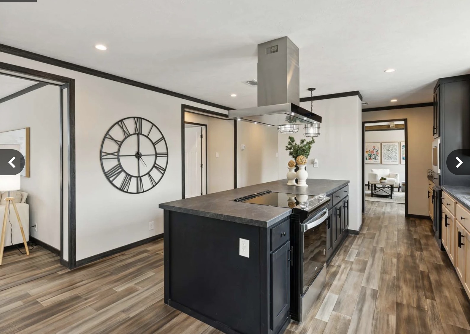 Modern kitchen with dark island and built-in stove, accented by a large wall clock and wooden floor. Adjacent room visible through doorway. Cozy ambiance.