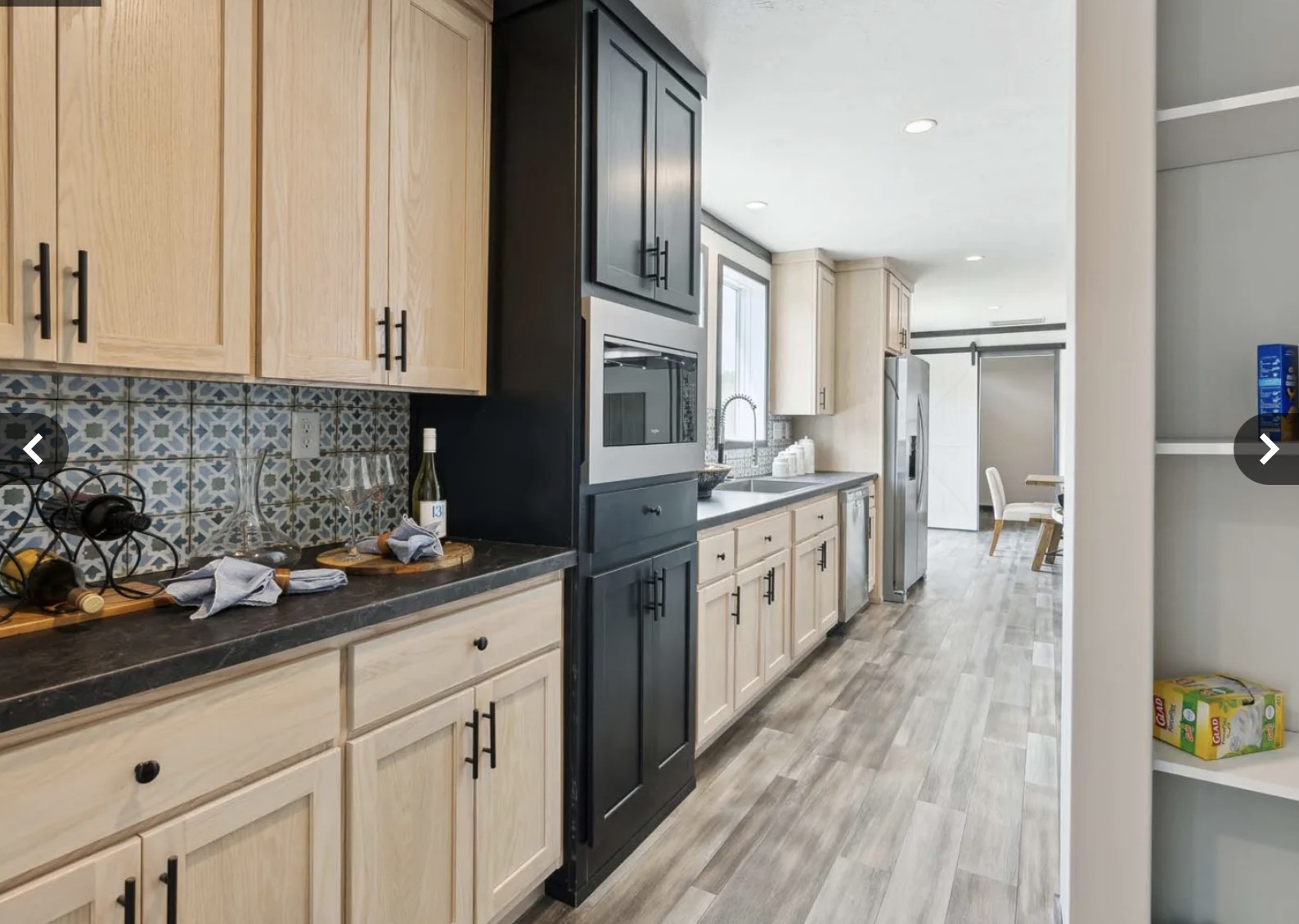 Modern kitchen with light wood cabinets, black countertops, and patterned backsplash. A wine bottle sits on the counter. Adjacent dining area visible.