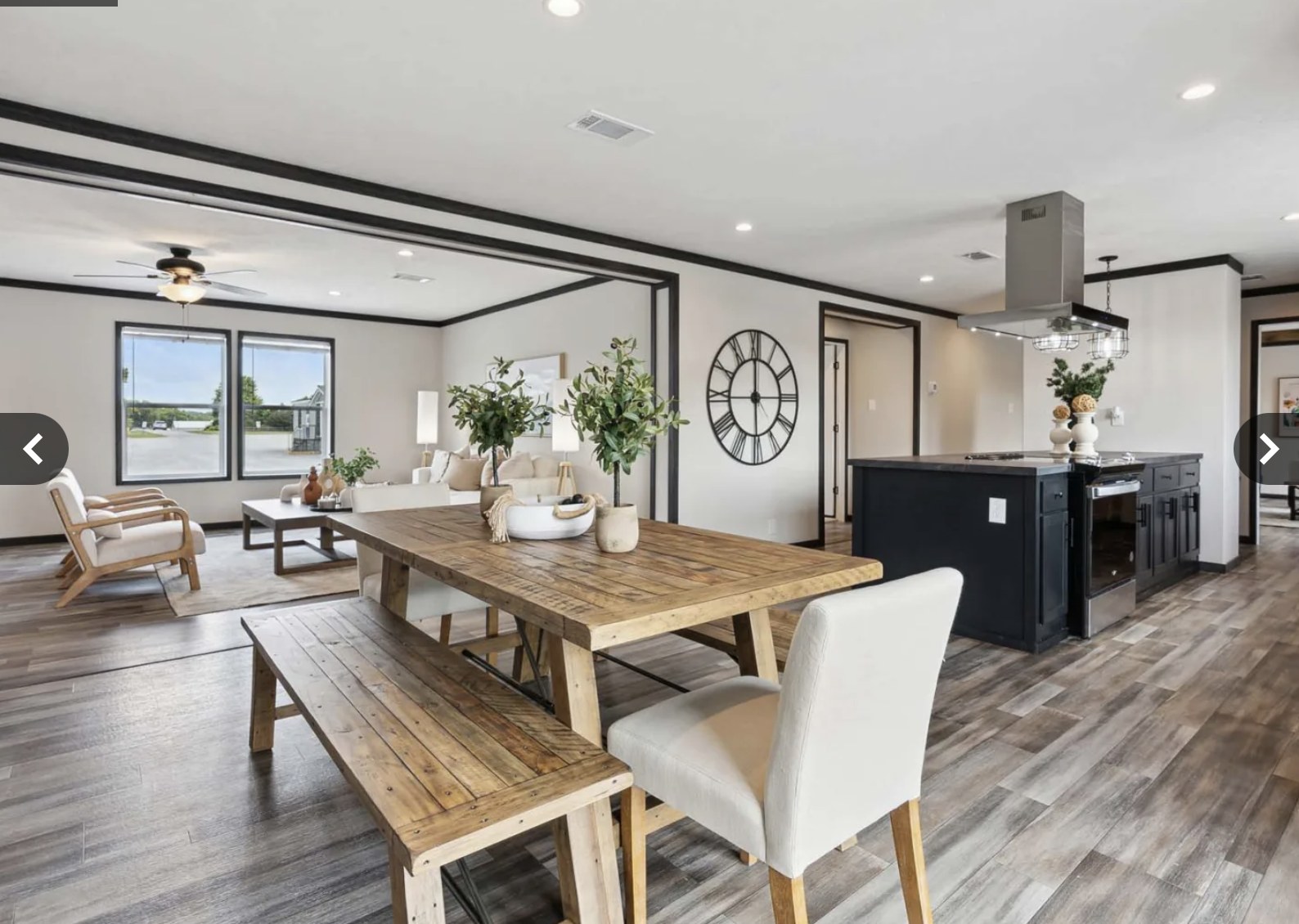 Modern open-concept living and dining area with wooden floors. Includes a rustic wooden table, white chairs, a cozy seating area, and a large wall clock.