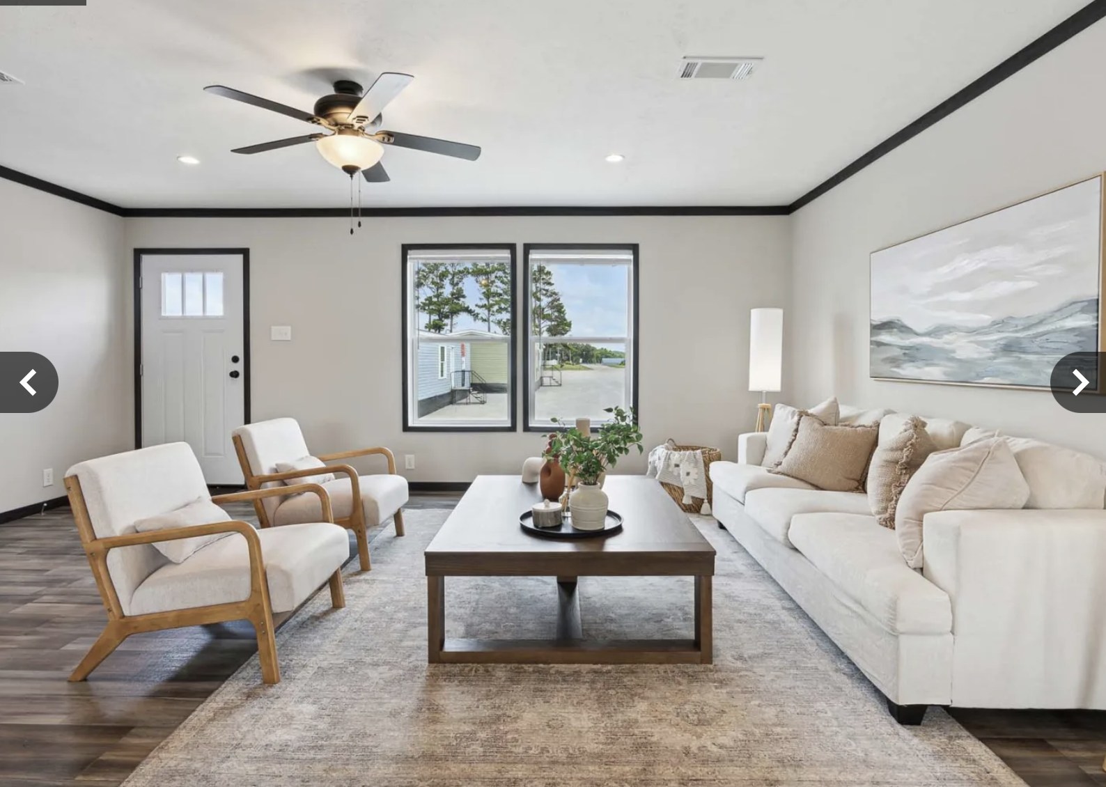 Modern living room with neutral tones, featuring a white sofa, two wooden armchairs, a coffee table, and large windows showing trees. Warm and inviting atmosphere.
